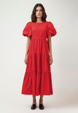 Žena stojaca, oblečená v červenom viacvrstvovom midi šate s nafúknutými krátkymi rukávmi a čiernych nazúvacích topánkach na bielom pozadí.