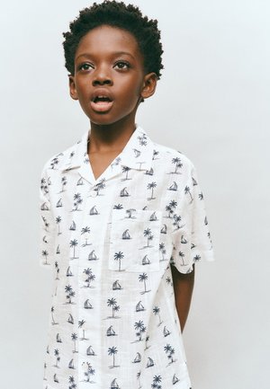 Chlapec s krátkymi kučeravými vlasmi, oblečený v bielej košeli s potlačou palm a plachetníc, pozerá sa dopredu s mierne otvorenými ústami.