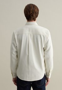 Witte langekranshemd met een gladde textuur, knoopsluiting, achterplooien en een gebogen zoom, gecombineerd met donkere spijkerbroek.