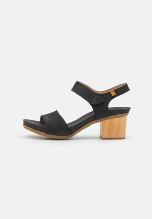 Sandale noire à bout ouvert avec bride de cheville réglable et talon bloc en bois sur fond blanc.