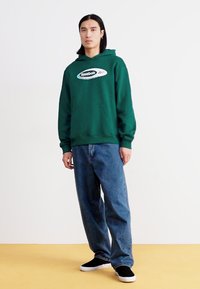 Hanorac verde cu buzunar frontal și logo alb, purtat cu blugi largi albaștri și pantofi slip-on negri, purtat de un model în picioare.