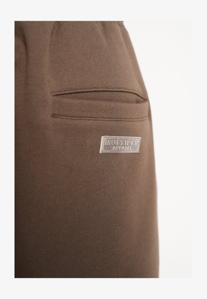 Braune Baumwoll-Jogginghose mit einer Seitentasche und einem reflektierenden gummiartigen Etikett mit der Aufschrift "MULTI-PAY APPAREL". Glatte Textur, taillierter Schnitt.