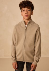 Maglione beige con zip, collo alto, trama lavorata a maglia e polsini a coste, indossato sopra una camicia bianca e abbinato a jeans scuri.