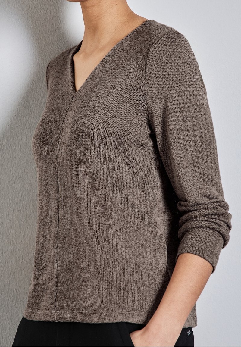 Femme portant un pull en maille marron à manches longues avec un col en V, debout devant un mur gris clair texturé.