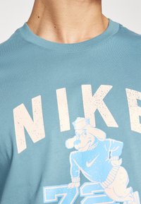 Lyseblå bomuldst-shirt med fed, hvid "NIKE" tekst og en tegneseriehund iført en sweater, med et legefuldt udtryk.