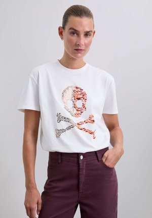 Camiseta blanca de algodón con un gráfico de calavera y tibias cruzadas colorido y texturizado. El modelo lleva pantalones burdeos oscuros, de pie con una mano en el bolsillo.