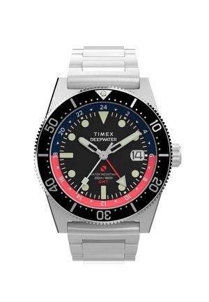Man in een zwart wetsuit past zijn nek aan en toont een polshorloge met een zwarte wijzerplaat en een meerkleurige bezel onder zacht blauw licht.