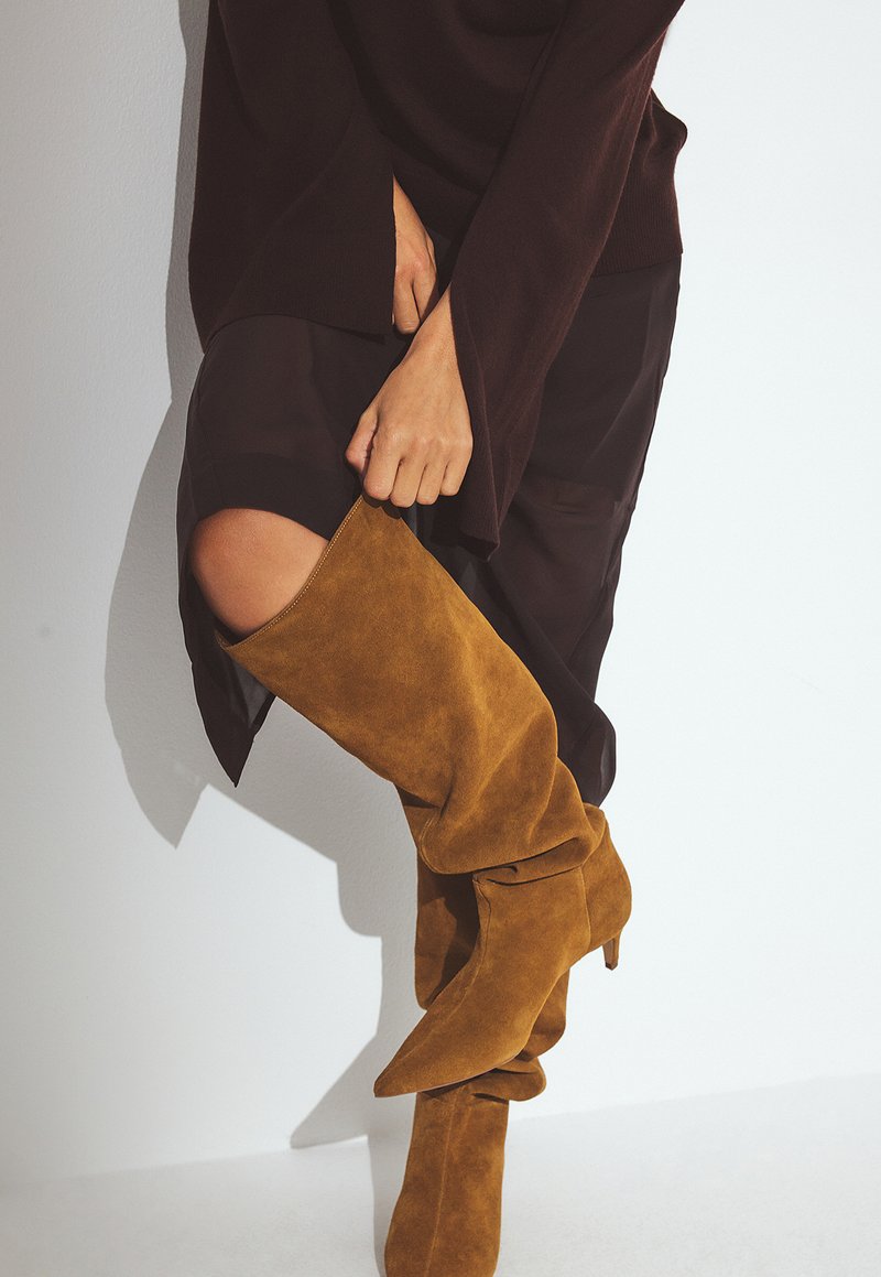 Bottes en daim moutarde montantes, avec un bout pointu et un design décontracté, associées à une tenue sombre et superposée comportant une fente sur la cuisse.