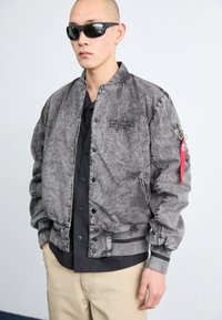 Gray bomber jacket with a textured finish, ribbed cuffs and hem, front buttons, side pockets, and a red tag accent on the sleeve.