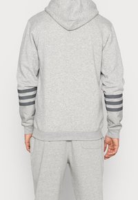 Osoba v světle šedé mikině s kapucí a odpovídajících kalhotách, se tmavě šedými a bílými pruhy na dolních rukávech, ukázaná zezadu.