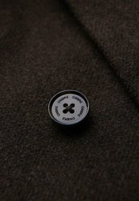 Tmavě hnědá látka s lesklým černým knoflíkem se čtyřmi otvory a slovem "OMBRE" opakujícím se kolem okraje.