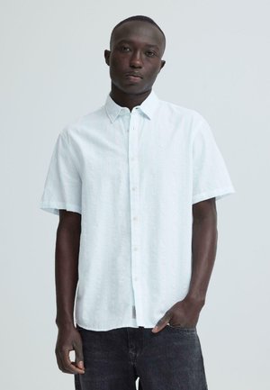 Jeune homme à la peau foncée portant une chemise blanche à manches courtes avec boutons et un jean noir, debout devant un fond clair uni.