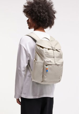 Beige batoh s horním klopovým víkem, nastavitelnými popruhy, bočními kapsami a dekorační visačkou s modrými, oranžovými a zelenými akcenty.
