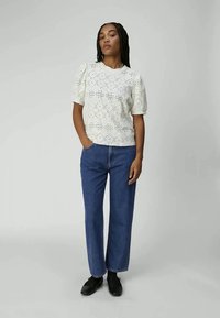 Woman standing with one hand in pocket, wearing white eyelet short-sleeve blouse, blue straight-leg jeans, and black shoes against plain background.