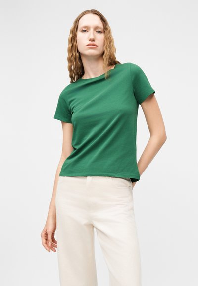 Femme aux cheveux châtain clair ondulés portant une chemise verte ajustée à manches courtes et un pantalon taille haute blanc cassé sur un fond uni.