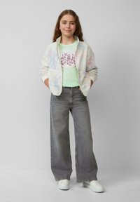Flauschige, pastellfarbene Zip-Jacke über einem hellgrünen Grafik-T-Shirt. Helle graue weit geschnittene Jeans und weiße Sneakers runden das Outfit ab.