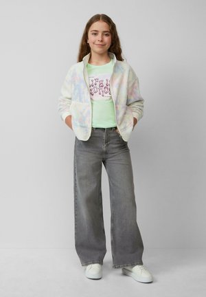 Een fuzzy, pastelgekleurde vest met rits over een lichtgroen t-shirt met opdruk. Lichtgrijze wijde spijkerbroek en witte sneakers maken de outfit compleet.