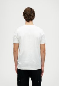 Muž s vlnitými hnědými vlasy, oblečený v jednoduchém bílém tričku s krátkým rukávem a černých cargo kalhotách, stojící zády k jednobarevnému pozadí.