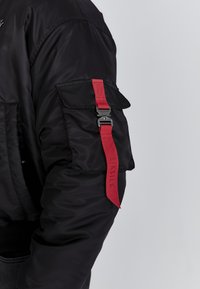Zwarte jas mouw met een klepzak en een rode stoffen band, bevestigd met een zwarte metalen gesp, met de opdruk "SIKSILK" in reliëf.