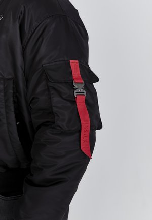 Black bomber jacket featuring a red strap with a clip on the left sleeve, smooth texture, and side pocket, showcasing a modern design.