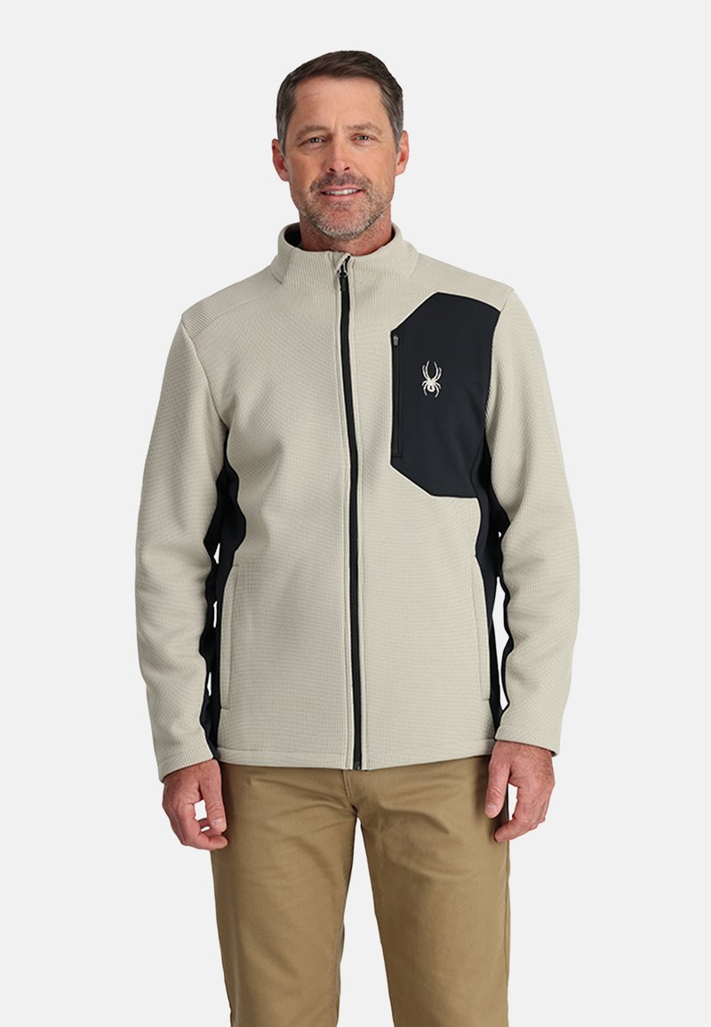 Veste en polaire gris clair avec une poche poitrine noire et une fermeture éclair. Présente une surface texturée et des accents de panneaux latéraux en noir.