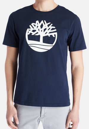 Person iført marineblå kortærmet t-shirt med hvidt stiliseret trælogo i midten af brystet, kombineret med lysegrå bukser.