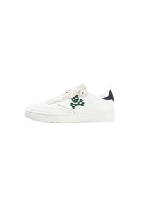 Zapatilla blanca de caña baja con acentos beiges, logo de calavera y tibias verdes en el costado, y detalle negro en el talón.