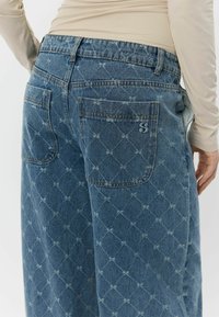 Blå denimjeans med et diamantmønster, fremhævede baglommer og broderet logo. Stoffet har et let falmet, tekstureret udseende.