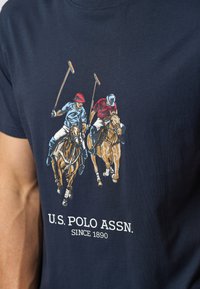 Laivyno spalvos medvilnės T-marškiniai su ryškiomis dviejų polo žaidėjų iliustracijomis ir tekstu "U.S. POLO ASSN. SINCE 1890" apačioje.