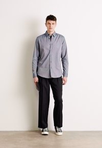 Gray button-up shirt, black trousers, and black-and-white sneakers. Model stands against a plain background, holding a clutch in one hand.
