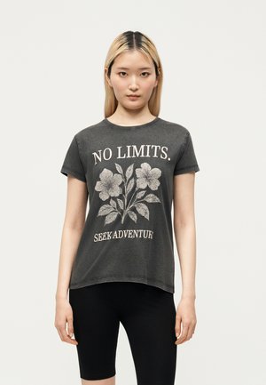 Ung kvinde med lige blondt hår iført en mørkegrå T-shirt med blomstermotiv og teksten "NO LIMITS. SEEK ADVENTURE" og sorte shorts.