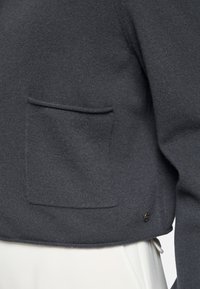 Dunkelgrauer Strickpullover mit einer kleinen aufgesetzten Tasche und einem metallischen Akzentdetail. Weiche Textur mit figurbetontem Design.