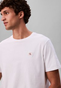 Bijela pamučna majica s okruglim izrezom, kratkim rukavima i malom crvenom oznakom CK logotipa na lijevoj strani prsa. Glatka tekstura.