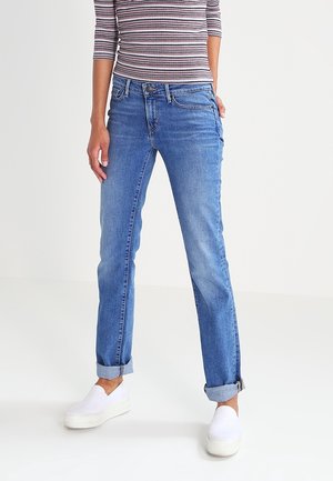 Personne portant un jean bleu retroussé, des chaussures blanches à enfiler et une chemise rayée à manches longues, debout devant un fond clair uni.