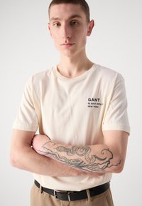 Beige katoenen t-shirt met korte mouwen, met zwarte geborduurde tekst: "GANT 32 EAST 39TH ST. NEW YORK." Model heeft een zichtbare tattoo op zijn arm.