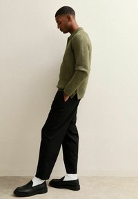 Suéter de punto verde oliva con un bolsillo frontal, pantalones negros ajustados, calcetines blancos y zapatos negros tipo slip-on. Diseño sencillo con acentos mínimos.