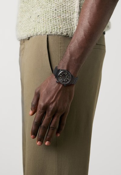 Black wristwatch with a round face, featuring gold hour markers and a textured black silicone band, worn on a hand beside an olive green pant.