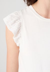 Top blanco de jersey con mangas de ojales con volantes. El top es suave con una silueta ajustada, que resalta el diseño detallado de las mangas.