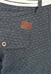 Par maramica tamno sive hlače s teksturiranim materijalom koji sadrži mali bijeli uzorak. Uključuje smešnu kožnu oznaku i drveni gumb kao detalj.