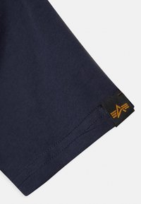 Tamno plava pamučna majica s kratkim rukavima, s crnom etiketom s zlatnim logotipom na rubu. Glatka tekstura i uredno šivanje.