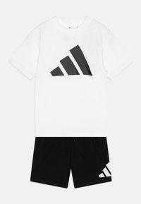 Camiseta de algodón blanca con un gran logotipo negro de Adidas. Combinada con pantalones cortos negros, que presentan un detalle de franja blanca en el costado.