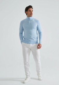 Pull bleu clair avec col zippé, manches longues et poignets côtelés, associé à un pantalon blanc et des baskets blanches sur un fond neutre.