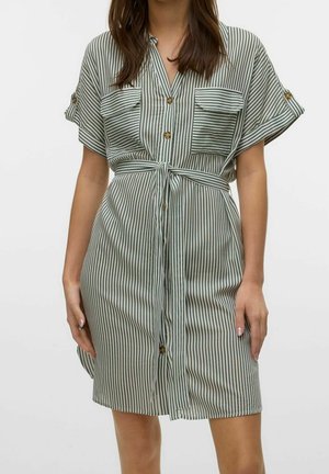 Femme portant une robe à manches courtes boutonnée avec des rayures verticales noires et blanches, des poches poitrine et une ceinture nouée à la taille.