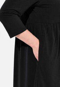 Mustapuku, jossa on rento leikkaus, kerätty vyötärö ja sivutasku. Valmistettu sileästä, joustavasta kankaasta, jossa on kolme neljännestä hihaa.