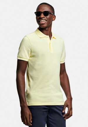 Uomo sorridente che indossa occhiali da sole tartarugati, polo giallo chiaro a maniche corte e pantaloni scuri, in piedi su sfondo bianco.