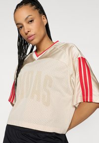 Jersey de malla beige con acentos rojos, que cuenta con mangas cortas, cuello en V y detalle de logo. La tela parece ligera y transpirable.