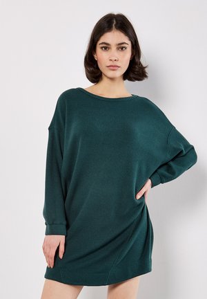 Vrouw met kort, donker haar draagt een oversized, gestructureerde, donkergroene jurk met lange mouwen, staand met één hand op de heup tegen een witte achtergrond.