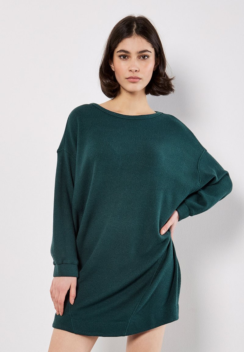 Vrouw met kort, donker haar draagt een oversized, gestructureerde, donkergroene jurk met lange mouwen, staand met één hand op de heup tegen een witte achtergrond.