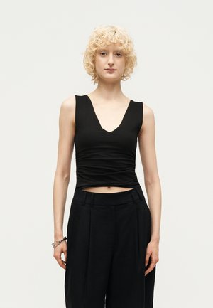 Jeune personne aux cheveux blonds courts et bouclés, portant un haut noir sans manches à col en V et un pantalon noir à jambes larges, sur un fond uni.