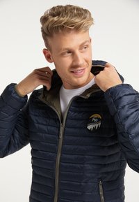 Giacca puffy blu navy con design trapuntato orizzontale, chiusura con zip, rivestimento verde e un patch rotondo con logo sul lato sinistro del petto.